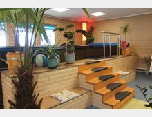 Innenraum eines Wellnessbereichs mit Holzverkleidungen und Pflanzen. Eine kleine Treppe mit Antirutschmatten führt zu einem erhöhten Bereich mit Geländer und Sitzkissen, umgeben von dekorativen Elementen wie Kübeln und einem aufblasbaren Ball.