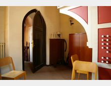 Ein Innenraum mit schlichten Holzmöbeln, einer roten Orgelkonsole mit Knöpfen, einem dunklen Spitzbogentürrahmen und einem angrenzenden Treppenbereich. Links ist ein Heizkörper sichtbar, rechts stehen Holzschränke und Reinigungsgeräte.