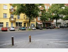 Straßenszene in einem städtischen Wohngebiet mit mehrstöckigen Gebäuden in gelber und brauner Farbe, Balkonen mit Blumenkästen und Schaufenstern im Erdgeschoss. Mehrere Autos sind am Straßenrand geparkt, und große Bäume stehen entlang des Gehwegs.