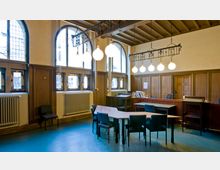 Ein historischer Innenraum mit Holztäfelung, hohen Fenstern mit Sprossenrahmen und mehreren Heizkörpern entlang der Wand. Ein zentraler Tisch mit Stühlen und eine Hängelampe mit kugelförmigen Glühbirnen dominieren den Raum, der von natürlichem und künstlichem Licht beleuchtet wird.