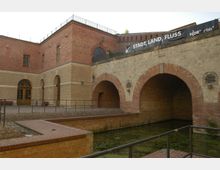 Backsteinbau mit historischen Bögen, einem kleinen Wassergraben im Vordergrund und einem Geländer. Auf der oberen Ebene des Gebäudes ist ein Banner mit der Aufschrift „Stadt, Land, Fluss“ sichtbar.