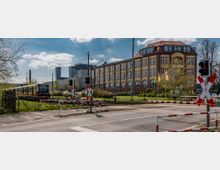 Bahnübergang mit Schranken und Signalleuchten, im Hintergrund ein gelbes Backsteingebäude mit großen Fenstern und rotem Dach, daneben moderne Industriegebäude und ein vorbeifahrender Zug auf den Gleisen. Im Vordergrund sind Straßenbegrenzungen und Grünflächen sichtbar, umgeben von Bäumen.