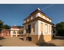 Historisches Gebäudeensemble mit barockem Stil, bestehend aus mehreren hellgelben und weißen Gebäuden mit dekorativen Fenstereinfassungen, rotem Walmdach und einem gepflasterten Vorplatz. Umgeben von grünen Bäumen unter einem klaren, blauen Himmel.