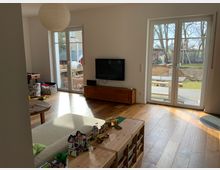 Wohnzimmer mit hellem Holzboden, einem niedrigen Holztisch, einem Sofa und Spielzeug, darunter Legobausteine. Große Fenster und eine Glastür führen zu einem Garten mit Holzterrasse und Bäumen im Hintergrund.