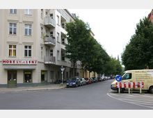 Straßenecke mit einem beige-farbenen Wohngebäude mit Balkonen, im Erdgeschoss ein Geschäft mit der Aufschrift „Möbel Eck Lenz“. Rechts eine Reihe von geparkten Autos entlang einer baumbestandenen Straße, im Vordergrund ein gelber Lieferwagen und eine Verkehrsinsel.