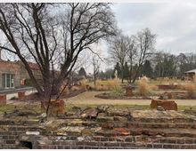 Ein Park mit kahlen Bäumen im Winter, Ziergräsern und gepflasterten Wegen. Im Vordergrund eine niedrige, teils zerfallene Backsteinmauer, im Hintergrund eine offene Rasenfläche und ein kleiner Holzpavillon.