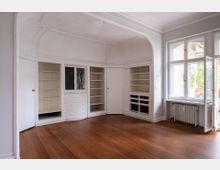 Heller, leerer Altbau-Raum mit hohen Decken, Stuckbögen und eingebauten weißen Schrank- und Regalflächen entlang der Wand. Rechts sorgen große Sprossenfenster neben einem Heizkörper für Tageslicht; der Boden besteht aus breiten Holzdielen.