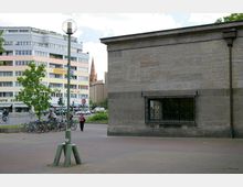 Städtischer Platz mit gepflastertem Boden, im Hintergrund ein modernes mehrstöckiges Wohn- und Geschäftshaus sowie der Turm einer Kirche. Im Vordergrund stehen Fahrräder an einem Fahrradständer, eine Straßenlaterne und ein Teil eines Gebäudes mit vergittertem Fenster sind sichtbar.