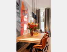 Moderner Essbereich mit langem Holztisch und braunen Lederstühlen, darüber eine Reihe schwarzer Pendelleuchten. An der Wand hängt ein großformatiges Wandbild, daneben bodentiefe Fenster mit grauen Vorhängen und Tageslicht.