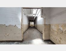 Langer, schmaler Flur eines verlassenen Gebäudes mit abblätternder Farbe an den Wänden und abgenutztem Boden. Die Decke ist hoch und zeigt freiliegende Rohre sowie Oberlichter, die natürliches Licht in den Raum lassen.