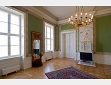 Ein eleganter Raum mit grünen Wänden, weißen dekorativen Türrahmen und hölzernem Parkettboden. Der Raum enthält einen großen Kronleuchter, einen verzierten Kachelofen, einen antiken Spiegel und einen orientalischen Teppich.