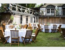 Ein Außenbereich mit rund angeordneten Tischen, die mit weißen Tischdecken bedeckt sind, umgeben von Holzstühlen auf einer Rasenfläche. Im Hintergrund ist ein rustikales Gebäude mit Holzverkleidung, großen Fenstern und einem oberen Balkon sichtbar.