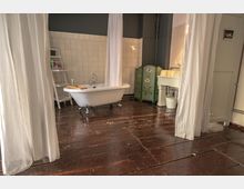 Ein Badezimmer mit einer freistehenden, weißen Badewanne auf Löwenfüßen, umgeben von weißen Vorhängen. Der Raum hat einen dunklen Holzboden, eine geflieste Wand, ein Waschbecken mit Unterschrank und einen bemalten Paravent mit Blumenmuster.