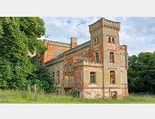 Verfallenes historisches Gebäude mit teilweise abblätterndem Putz und freiliegenden Ziegelsteinen, einem Eckturm und Rundbögen als Zierelementen. Das Gebäude ist von Grünflächen und Bäumen umgeben, teilweise eingezäunt.