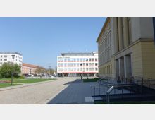 Städtischer Platz mit gepflastertem Weg, umgeben von modernen und historischen Gebäuden, darunter ein gelbes Gebäude mit Säulen rechts im Bild. In der Mitte steht ein weißes Gebäude mit der Aufschrift „Linden-Zentrum Bibliothek.“