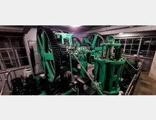 Industrieraum mit einer historischen grünen Maschine, die große Zahnräder, Hebel und Metallrohre umfasst. Im Hintergrund sind große Fenster und metallene Wände sichtbar.