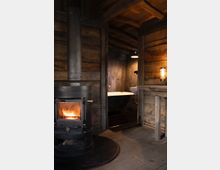 Gemütlicher Innenraum aus Holz mit einem schwarzen Kaminofen auf einer Metallplatte im Vordergrund, der ein sichtbares Feuer enthält. Im Hintergrund führt eine Tür zu einem Bad mit einer freistehenden Badewanne, schummriger Beleuchtung und Betonwänden.