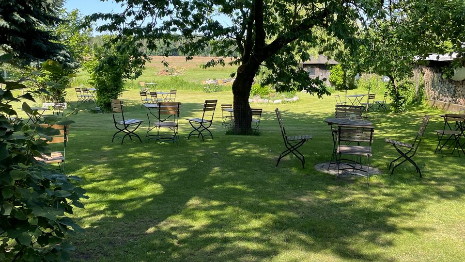 Große, gepflegte Rasenfläche mit schattenspendendem Baum in der Mitte und mehreren verstreut stehenden Metall-Gartentischen und -stühlen. Im Vordergrund blühen pinke Blumen, im Hintergrund sind Felder und einige niedrige Gebäude am Rand des Gartens zu sehen.