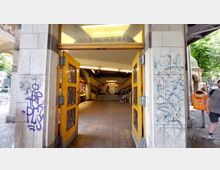 Eingang zu einer U-Bahn-Station mit offenen gelben Holztüren, gefliesten Wänden und Treppen im Inneren. Die Außenwände sind mit Graffiti beschmiert, und rechts steht ein orangefarbener Abfalleimer auf einem Gehweg mit Bäumen im Hintergrund.