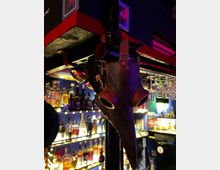 Eine Bar mit Regalen voller Flaschen und gläsernem Barzubehör im Hintergrund; im Vordergrund hängt eine dekorative schwarze Pestmaske mit auffälligem Schnabel und angenieten Details an einer Metallstange.