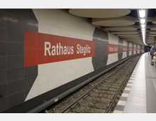 U-Bahn-Station mit dem Schriftzug „Rathaus Steglitz“ auf einer Wand aus grauen und weißen Fliesen, unterbrochen durch rote Streifen. Im Vordergrund befinden sich Gleise, und der Bahnsteig ist mit einem Fliesenmuster gestaltet.