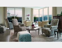 Ein moderner, heller Wohnraum mit Polstersesseln in neutralen Farbtönen und geometrischen Mustern, kleinen runden Tischen mit Dekorationen sowie großflächigen Fensterfronten, die auf einen Balkon mit Pflanzen und Blick ins Freie führen. Gardinen rahmen die Fenster, und im Hintergrund steht eine dunkelbraune Vitrine.