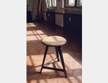 Ein einfacher Holzstuhl mit drei Metallbeinen steht in einem Raum mit großflächigen Fenstern, die Sonnenlicht auf den hellbraunen Boden werfen. An der Wand befinden sich Heizkörper und Fensterbänke.