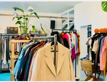 Ein Secondhand-Laden mit Kleidungsstücken auf Kleiderstangen, darunter Mäntel, Hemden und bunte Oberteile. Im Hintergrund sind ein Regal mit Zimmerpflanzen, eine Diskokugel an der Decke und eine Stereoanlage zu sehen.