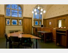 Raum mit historischer Holzvertäfelung, großen Bogenfenstern mit Gitterverglasung und einem zentralen Kronleuchter. Die Einrichtung umfasst Schreibtische, Stühle und einen Empfangstresen, gestaltet in dunklem Holz.