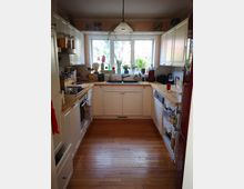 Helle Küche mit weißen Einbauschränken, Holzarbeitsplatten und einem Spülbecken unter einem großen Fenster. Der Raum verfügt über einen Holzboden, zahlreiche Küchengeräte und dekorative Pflanzen auf der Fensterbank.