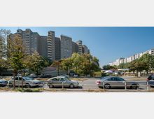 Wohngebiet mit einem großen, mehrstöckigen Gebäude im Hintergrund, dem "Gropiushaus", flankiert von weiteren Wohnblocks. Im Vordergrund ist eine Straße mit geparkten Autos, Bäumen und einer kleinen Grünfläche zu sehen.