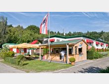Kleiner Kiosk aus Holz mit grünem Dach und Verkaufsfenster, umgeben von einer Rasenfläche mit Sitzmöglichkeiten unter gelben Sonnenschirmen. Im Hintergrund ein weißes Gebäude mit roten Markisen und ein angrenzendes Waldgebiet.