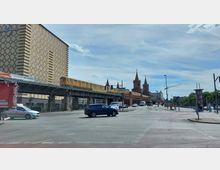Straßenszene in Berlin mit der Oberbaumbrücke im Hintergrund. Ein gelber U-Bahn-Zug fährt auf der erhöhten Bahntrasse entlang, rechts sind die charakteristischen Türme der Brücke zu sehen; links ein großes Ziegelgebäude mit der Aufschrift "Universal".
