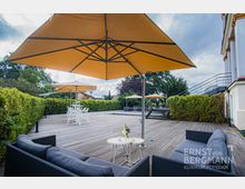 Eine Holzterrasse mit Sitzbereichen, grauen Sofas und runden Tischen, geschützt durch große gelbe Sonnenschirme. Der Bereich ist von hohen grünen Hecken umgeben, mit Treppen, die zu einer höheren Terrasse führen, und einem Teil eines weißen Gebäudes am rechten Rand.