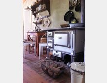 Eine rustikale Küche mit einem weißen, altmodischen Holzofen auf einem Ziegelboden, daneben ein Drahtkorb voller Holzscheite. Im Hintergrund ein Holztisch mit Stühlen, ein Wandregal mit Geschirr und Kochutensilien sowie an der Wand hängende Pfannen und Küchenutensilien.