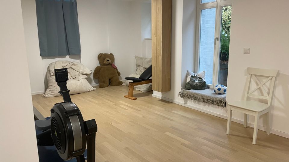 Heller Innenraum mit hellem Holzboden und weißen Wänden, eingerichtet mit einem Ruderergometer, einer flachen Trainingsbank und Sitzkissen. An der rechten Seite befindet sich ein großes Fenster mit Fensterbank-Sitzplatz sowie ein hoher Holzschrank und ein Stuhl.