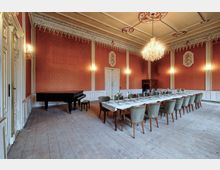 Großer historischer Festsaal mit hohen rot gemusterten Wänden, Stuckfriesen und einem Kristallleuchter an der Decke. In der Mitte steht eine lange gedeckte Tafel mit Polsterstühlen; links befindet sich ein schwarzer Flügel auf einem breiten Holzdielenboden.