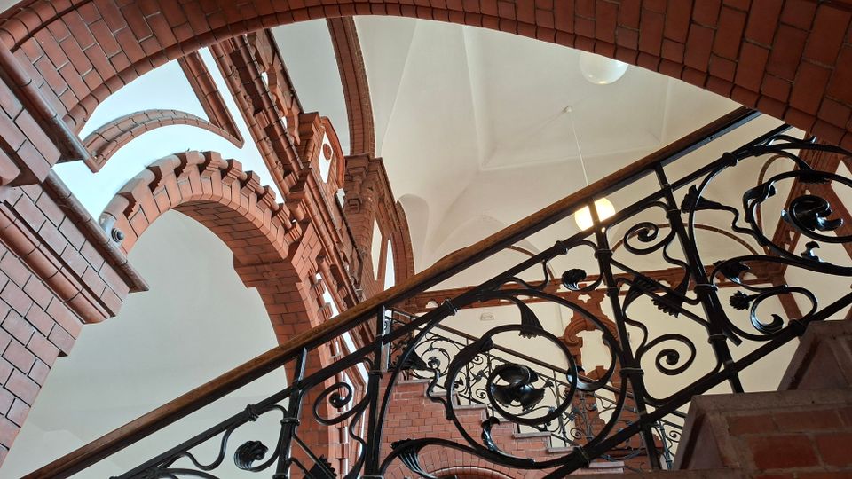 Innenansicht eines Treppenhauses mit roten Backsteinbögen und weißen Gewölbedecken. Eine geschwungene Treppe mit schmiedeeisernem Geländer und Holzhandlauf verläuft diagonal durch das Bild.