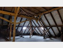 Dachboden mit sichtbarer Holzkonstruktion und Schrägdach, das mit weißer Dämmung ausgekleidet ist. Zwei kleine Fenster lassen Tageslicht in den Raum, der unbehandelte Böden und freiliegende Ziegelwände aufweist.