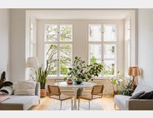 Heller Wohnraum mit großen Sprossenfenstern, die auf Bäume und eine gegenüberliegende Fassade blicken. Zentral steht ein runder Tisch mit zwei Stühlen, umgeben von Pflanzen, Sofas und Stehlampen auf Parkettboden.