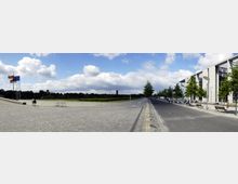 Weitläufiger, gepflasterter Platz mit Flaggenmasten auf der linken Seite, flankiert von einer Rasenfläche und Bäumen im Hintergrund; rechts ist eine moderne Gebäudefassade mit gläsernen Elementen und einer Baumreihe entlang einer Straße zu sehen. Der Himmel ist teils bewölkt.