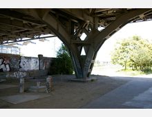 Ansicht unter einer stählernen Brückenkonstruktion mit großen, verzweigten Stützen. Im Hintergrund eine Backsteinmauer, die mit Graffiti bedeckt ist, sowie ein Grünbereich mit Bäumen im rechten Bildbereich.