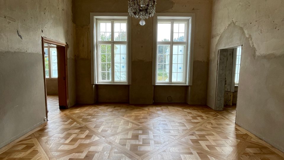 Großer, leerer Innenraum eines Altbaus mit hellem Parkett im geometrischen Muster, hohen Decken mit Stuckrosette und einem hängenden Kronleuchter. An der Stirnwand stehen zwei hohe Sprossenfenster; seitlich führen offene Durchgänge in weitere Räume, die Wände sind teils unverputzt und renovierungsbedürftig.