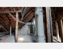 Dachboden eines alten Gebäudes mit sichtbaren Holzbalken, einer offenen Holzleiter, einem massiven Betonpfeiler und freiliegenden Rohrleitungen. Der Boden besteht aus Ziegeln, und an der Rückwand ist eine Tür erkennbar.