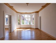 Helles Zimmer in einem Altbau mit geprägten Stuckverzierungen an der Decke, großen Sprossenfenstern und hellem Parkettboden. Ein Durchgang führt in ein angrenzendes Zimmer, von dem ebenfalls Fenster sichtbar sind.