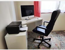 Ein einfach eingerichtetes Homeoffice mit weißem Schreibtisch, auf dem ein Monitor, eine Tastatur und eine Maus stehen. Daneben ein schwarzer Drucker auf einem Schrank; im Vordergrund ein schwarzer Bürostuhl und im Hintergrund ein Fenster mit roten Vorhängen.