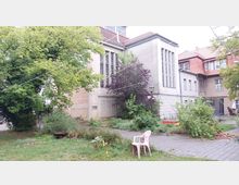 Außenbereich eines Gebäudekomplexes mit grauer und beiger Fassade, großen Fenstern und einem roten Ziegeldach. Der Garten ist von Bäumen, Büschen und Rasenflächen geprägt, mit einem gepflasterten Weg, einem rosa Plastikstuhl und einem Holzgestell im Hintergrund.