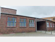 Backsteingebäude mit großen Fenstern, deren Rahmen in Rot und Blau gestrichen sind. Rechts befindet sich ein überdachter Durchgang, der von Pfeilern gestützt wird, und der Boden vor dem Gebäude ist mit Pflastersteinen bedeckt.