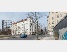 Straßenszene in einer Stadt mit mehrstöckigen Altbauwohngebäuden, Ampeln und geparkten Autos. Im linken Hintergrund ist ein großer Gasometer sichtbar, während rechts eine Mauer mit Graffiti bemalt ist.
