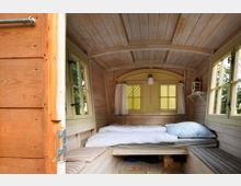 Innenansicht eines kleinen Holzwohnwagens mit rustikalem Interieur. Der Raum enthält ein Bett mit weißer Bettwäsche, Holzwände und -decke, gelbe Fensterrahmen mit Vorhängen sowie Sitzbänke mit Polstern an den Seiten.